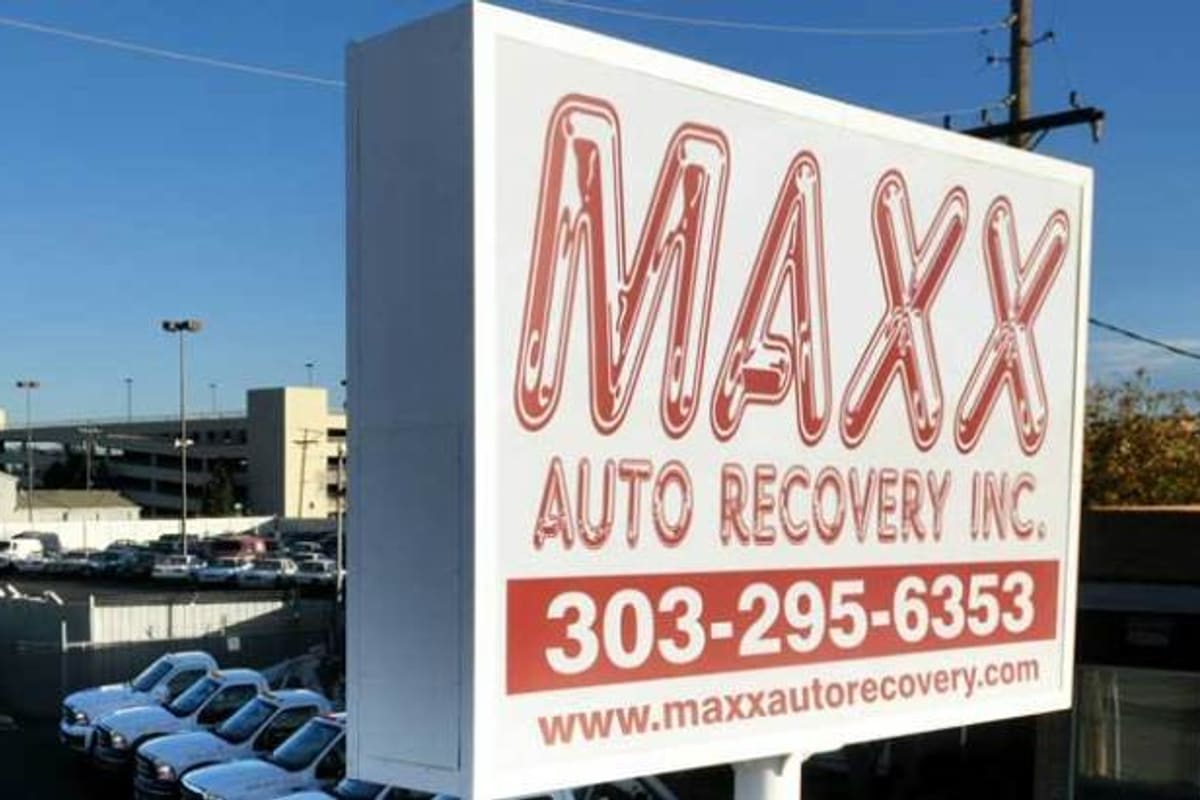 Maxx Auto Recovery: Large roadside sign beside a fenced lot filled with white tow trucks and parked vehicles under a blue sky