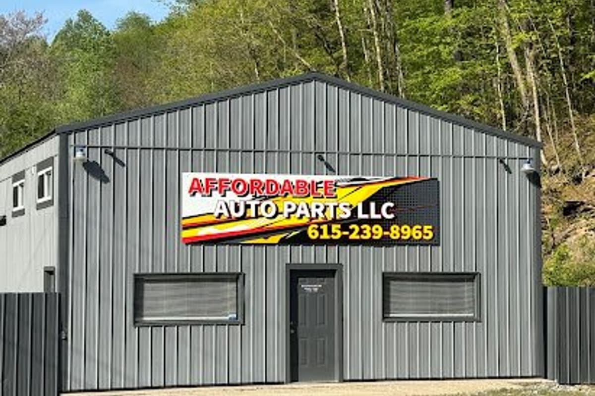 Affordable Auto Parts: Gray metal building with a large red, yellow and black sign above a dark entrance door, with wooded hillside behind