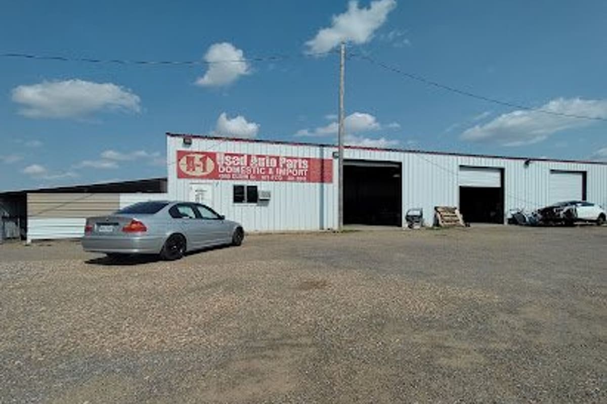 Le's Used Auto Parts & Used Cars: Metal shop building with red used auto parts sign, open bay doors and a gravel lot with a silver sedan parked in front