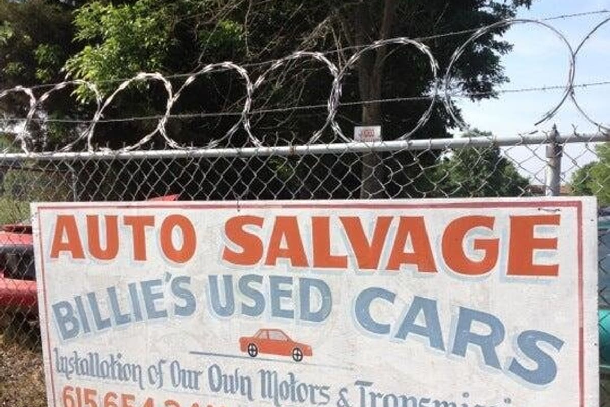 Billie's Used Cars & Repairs: Hand-painted auto salvage sign on a chain-link fence topped with razor wire, with entrance label and phone numbers