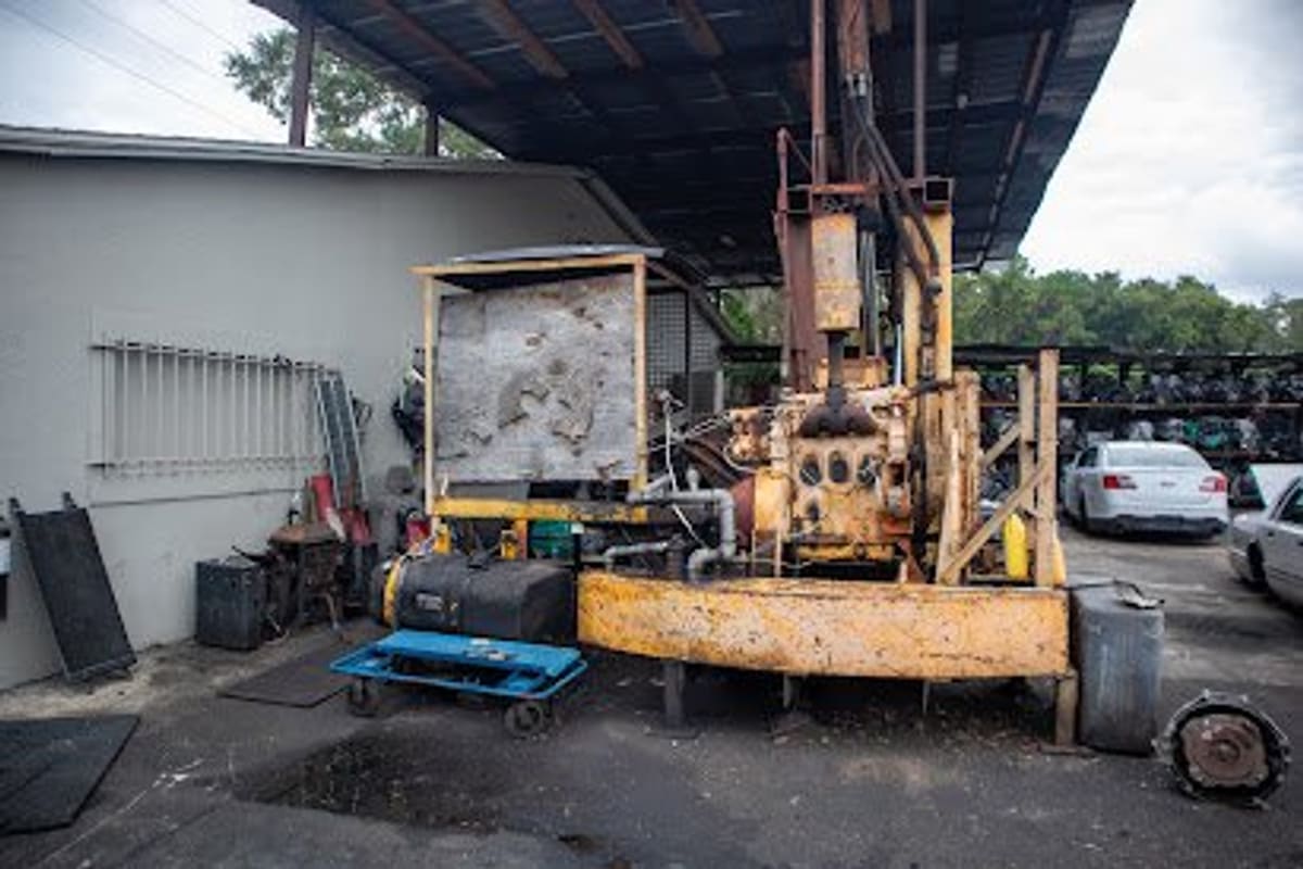 A&K Auto Recycling: Rusty yellow industrial machine under a covered area beside a gray building, with cars and stacked parts in the yard behind
