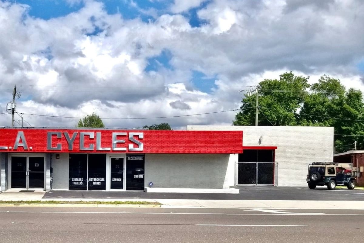 FL Sunshine MC Parts: Single-story motorcycle shop with a red brick sign band, glass doors and a paved parking area in front
