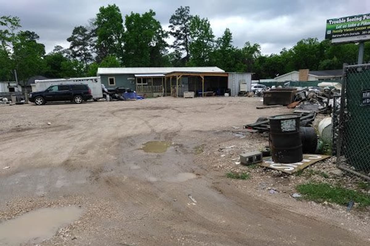 Uncle G Auto Recycling: Gravel salvage yard with a small shop building, scrap piles, a barrel and parked vehicles under a cloudy sky