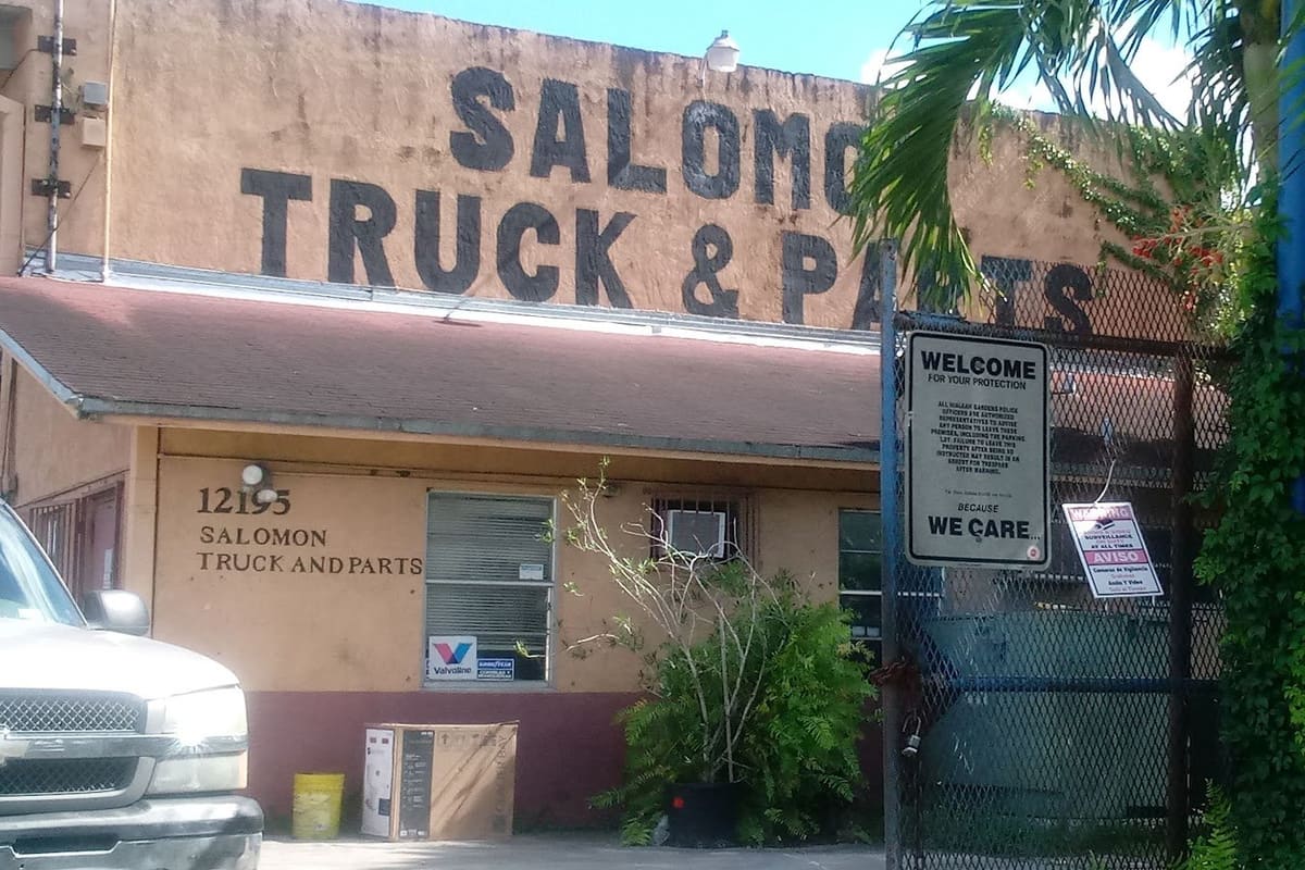 Salomon Truck Parts and Export: Weathered tan building with large painted lettering reading Truck & Parts, a chain-link gate and a street number beside the door