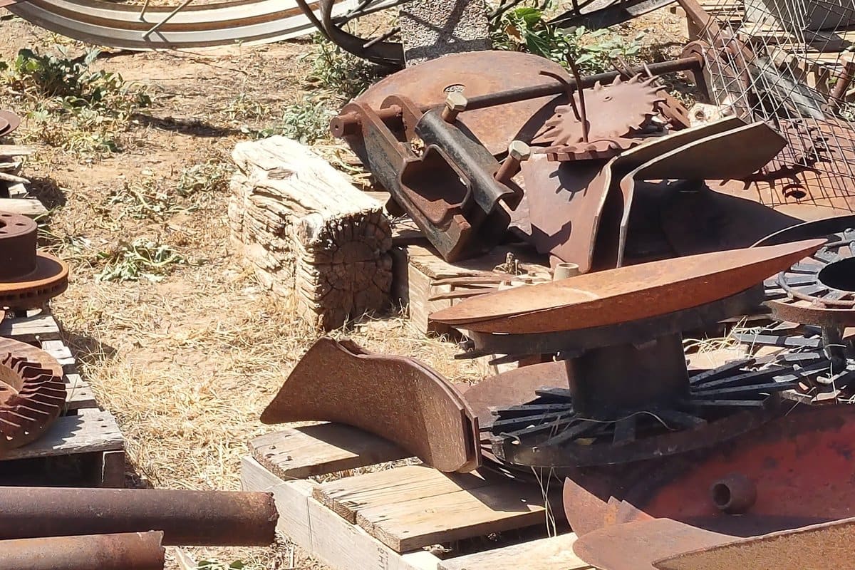 Big R Surplus & Salvage: Rusty antique wagon wheels and scrap metal parts piled on dirt inside a chain-link fence beside a road