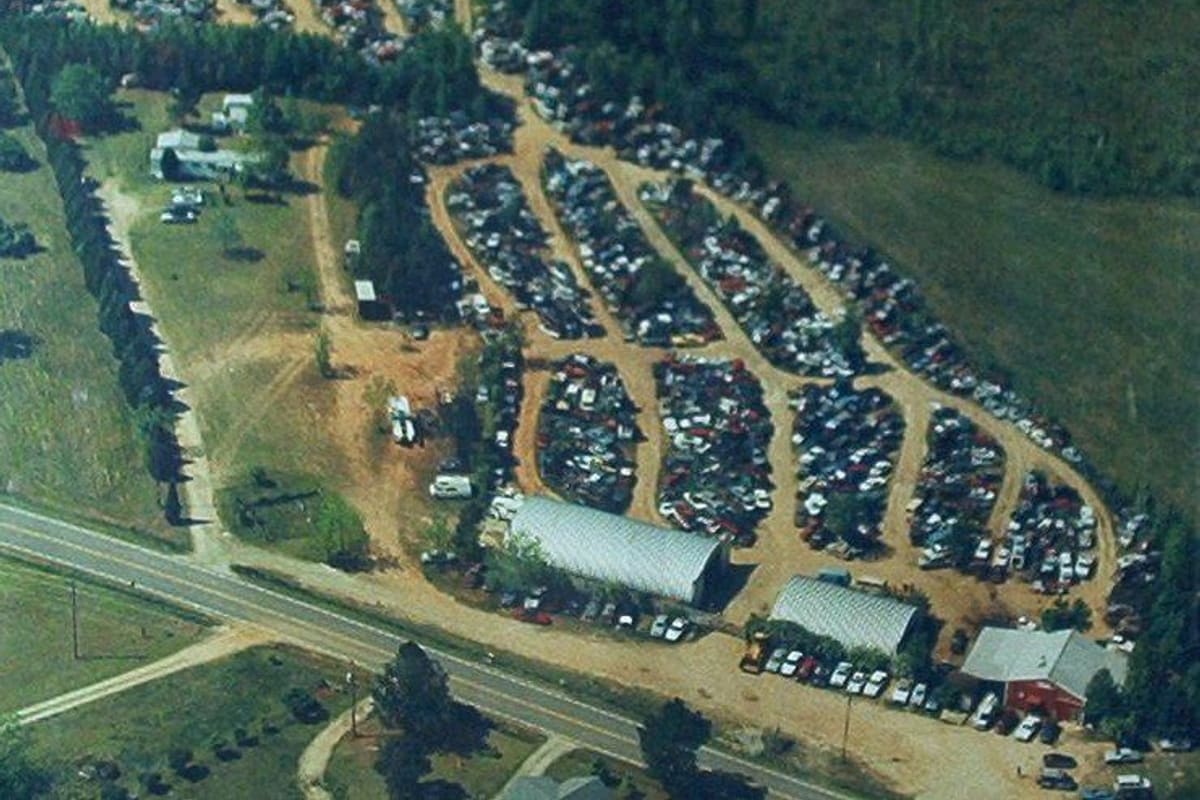 Johnston's Used Auto Parts: Aerial view of a salvage yard with rows of vehicles, metal-roofed buildings, and a dirt lot beside a highway