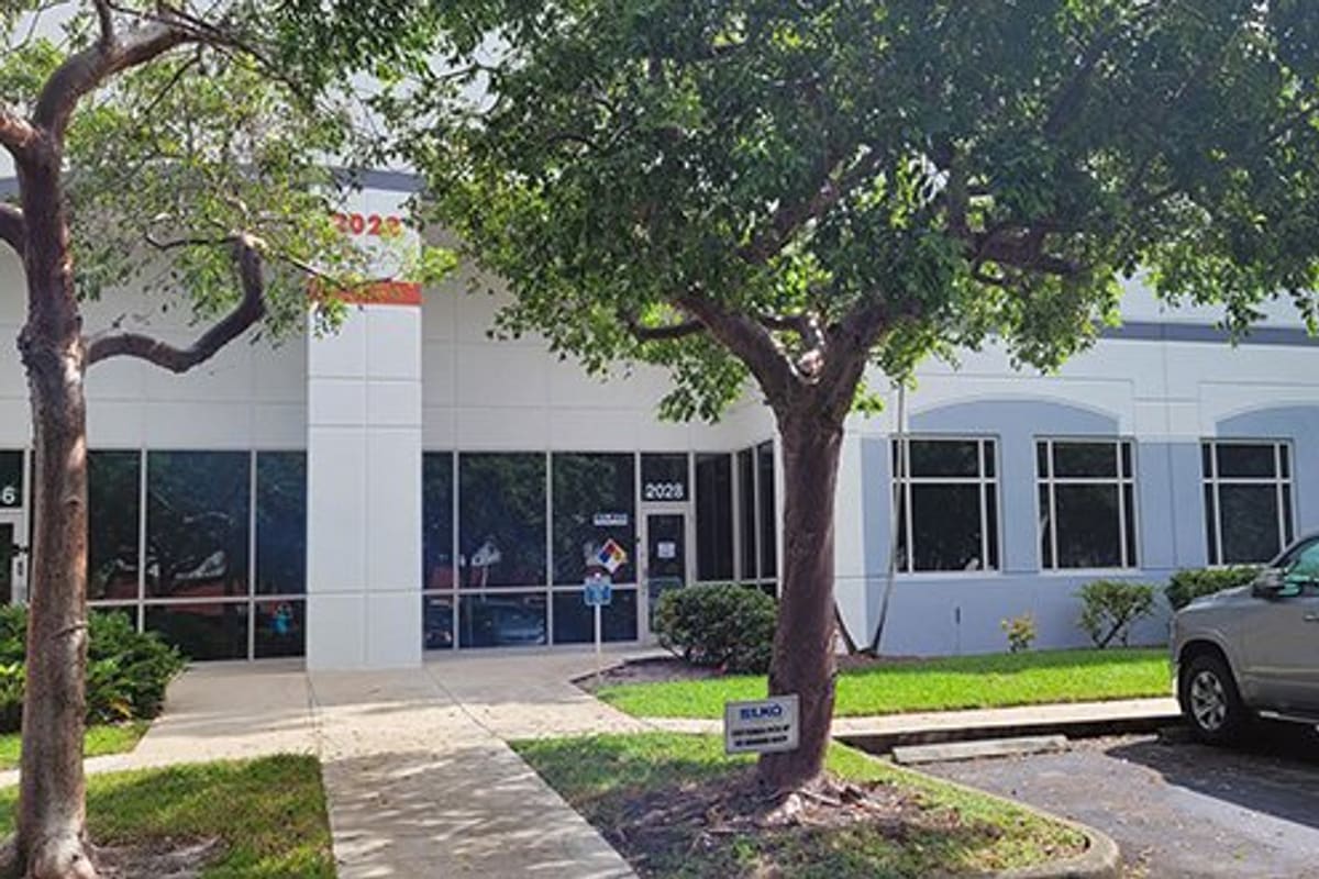 LKQ Melbourne - Pompano Beach: White commercial building with glass storefront entrance, trees, landscaped walkway, and pickup truck parked nearby