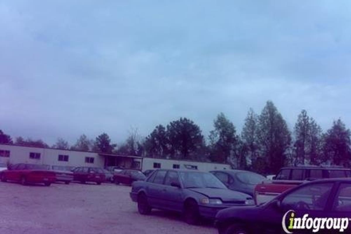 Kavita: Gravel lot with rows of parked used cars in front of a low building and trees under a cloudy sky