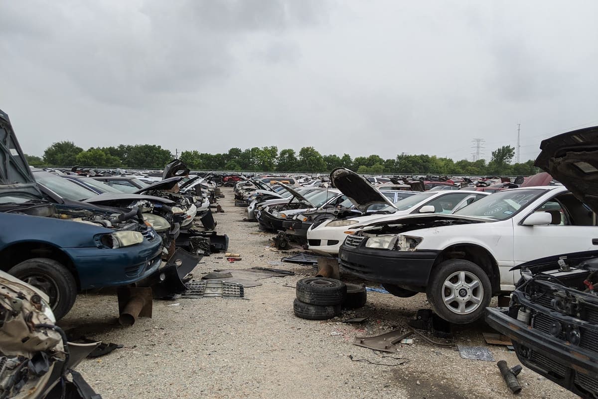 Pick Your Part - Houston SW: Rows of salvage cars with open hoods lined up along a gravel aisle in a self-service junkyard under a cloudy sky