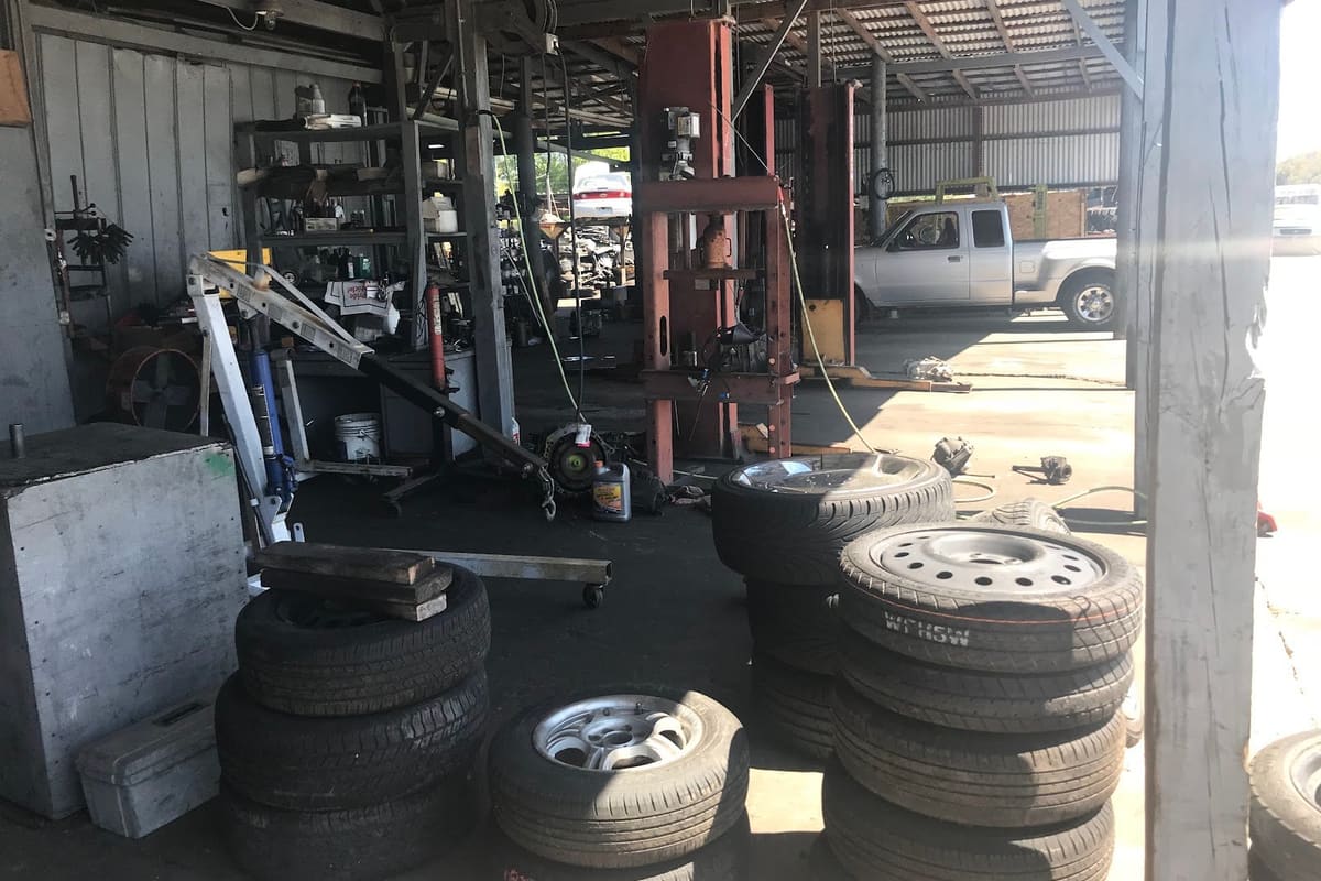 Saint Cloud Auto Salvage: Open-air covered repair area with stacked tires, a hydraulic press, an engine hoist and a pickup truck parked beyond