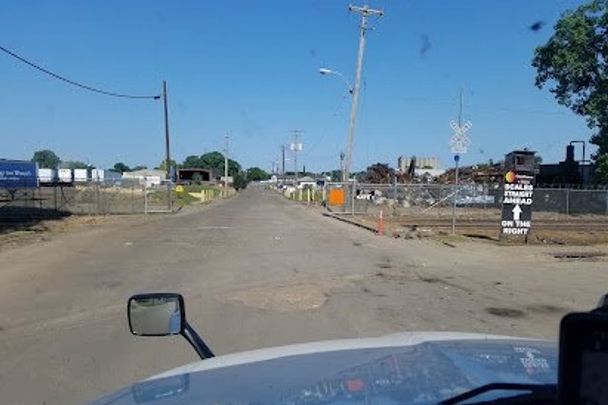 Omnisource: View from a truck cab down a paved industrial road toward a scrapyard, with a sign reading scales straight ahead on the right