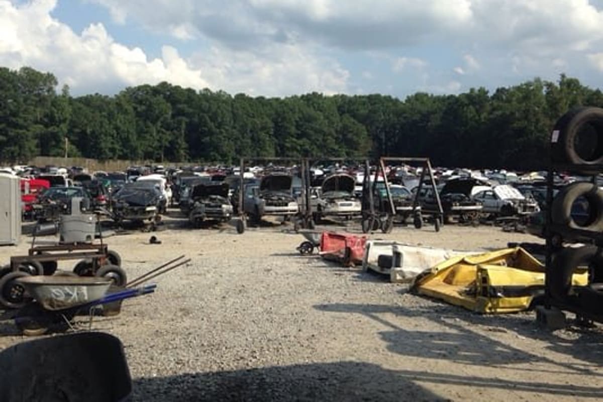 Pick Your Part - Raleigh: Gravel salvage yard with rows of junk cars on stands, tire racks and wheelbarrows under a cloudy sky