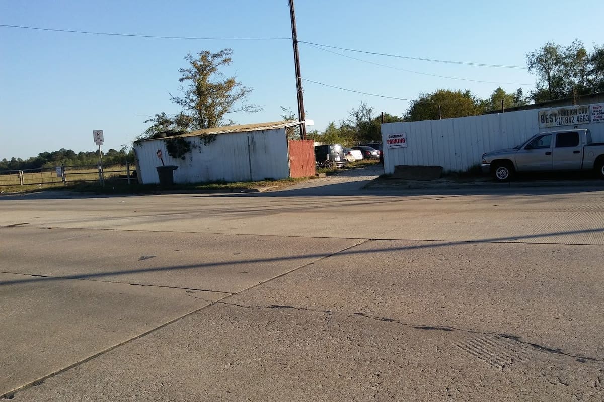 Best Imports Salvage: Wide daytime view across a concrete road to a salvage lot with a corrugated metal fence, customer parking sign and a pickup truck