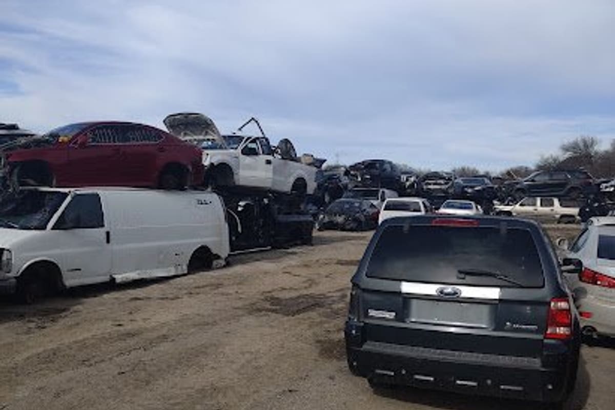 Hernandez Auto Salvage: Wide view of a salvage yard with stacked wrecked cars, vans and trucks on a dirt lot under a partly cloudy sky