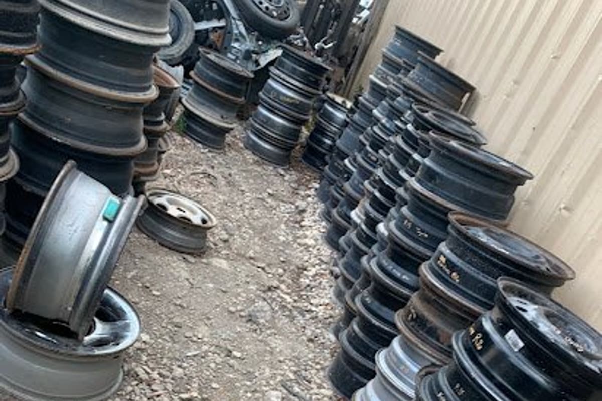 Parkway Used Auto Parts: Stacks of used steel and alloy wheel rims on gravel ground beside a metal building wall in a salvage yard