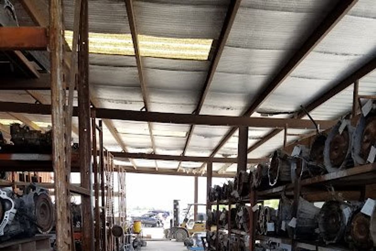 Assured Auto Parts: Covered aisle lined with metal shelving racks holding rows of used transmissions, with a forklift and cars in the distance