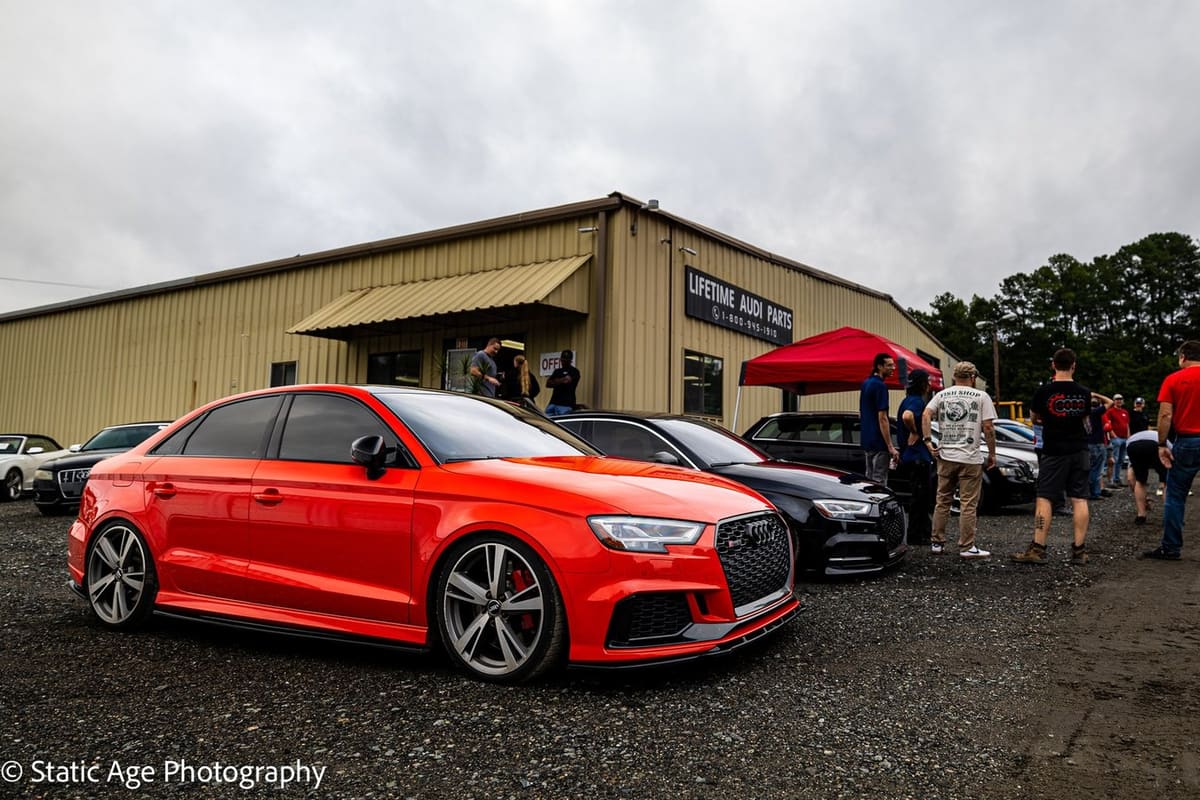 Lifetime Audi Parts: Metal warehouse building with an auto parts sign, a red Audi sedan and other cars parked on gravel as people gather