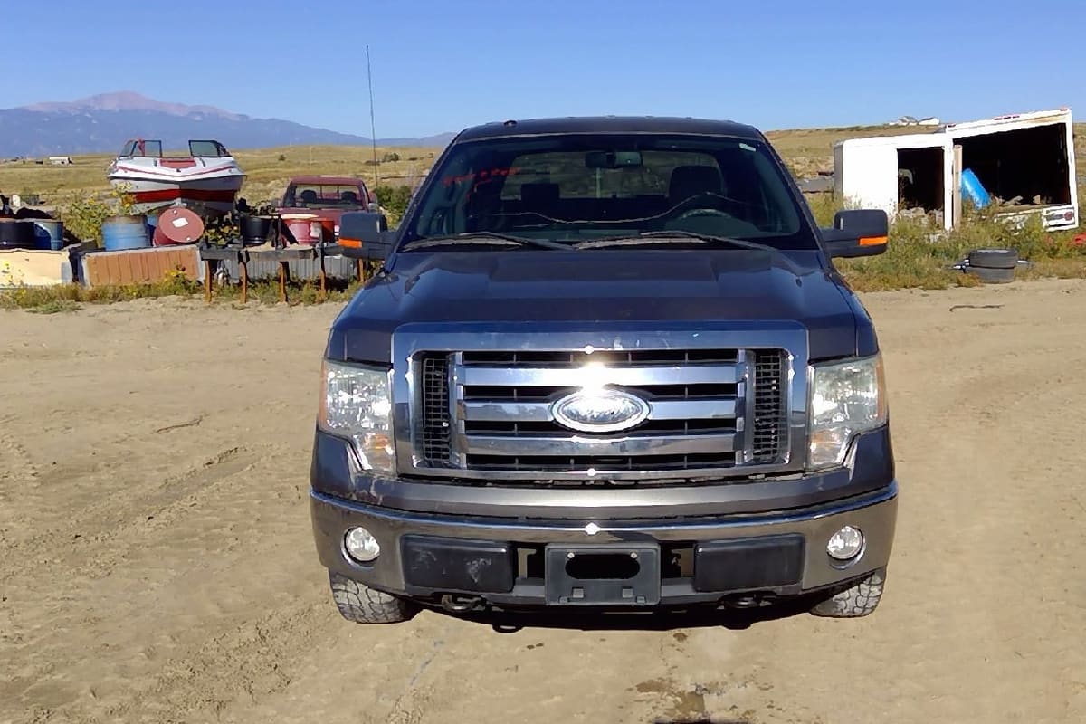 American & Import Auto Recyclers: Gray pickup truck parked on a dirt lot with a boat, trailers and scrap items and a mountain in the background