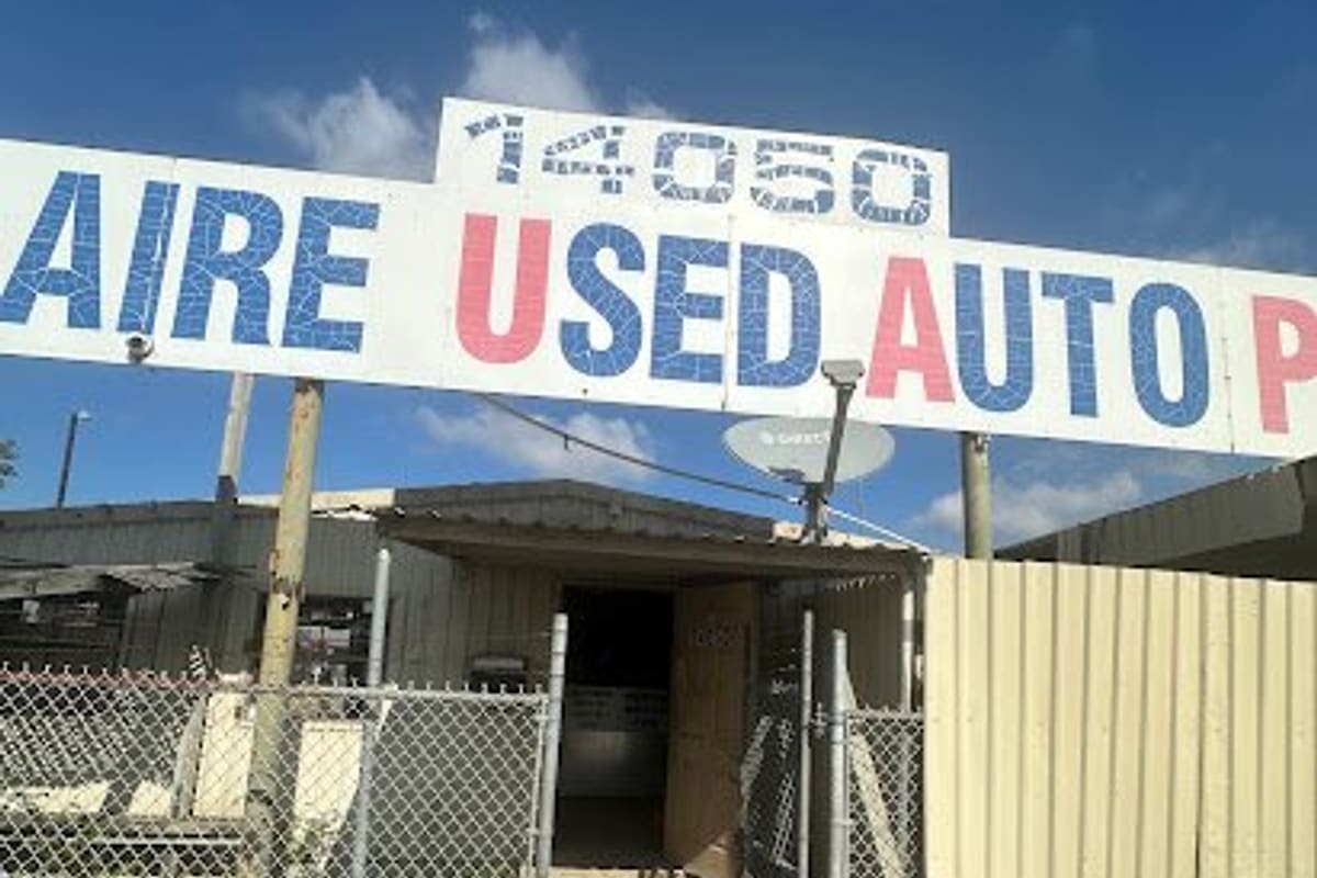 Bellaire Used Auto Parts: Large hand-lettered sign over a chain-link gate and walkway leading to a small metal office building under a blue sky