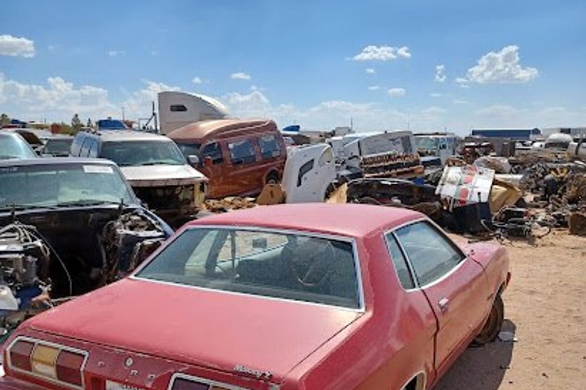 El Todo Yonke Yard: Red vintage coupe parked in a dirt salvage yard with rows of wrecked vehicles and parts under a blue sky