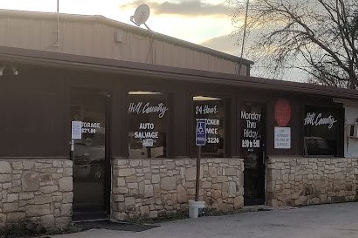 Hill Country Auto Salvage: Stone-fronted building with brown trim, storefront windows lettered with auto salvage and hours, and a gravel lot in front