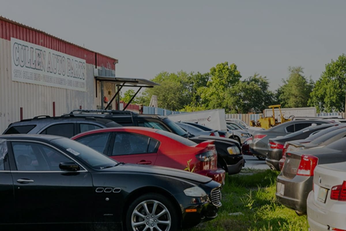 Parts R US: Rows of used cars parked on grass beside a metal warehouse building with a painted sign on a sunny day