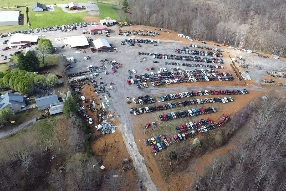 Smith's(john) Auto Truck Salvage: Aerial view of a large salvage yard with long rows of vehicles on dirt and gravel, surrounded by woods and several buildings