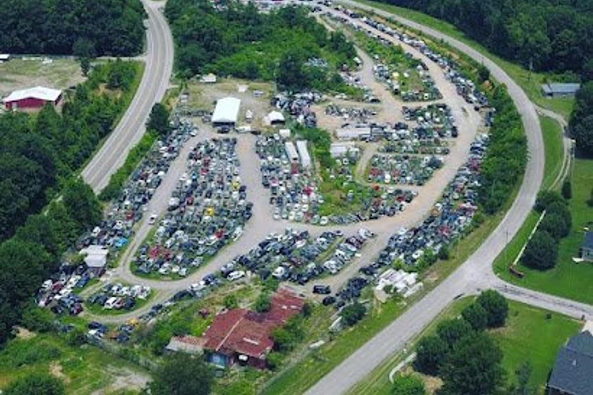 Woodbury Auto Salvage and Recycling: Aerial view of a large salvage yard with rows of vehicles along a curving road, surrounded by forested hills