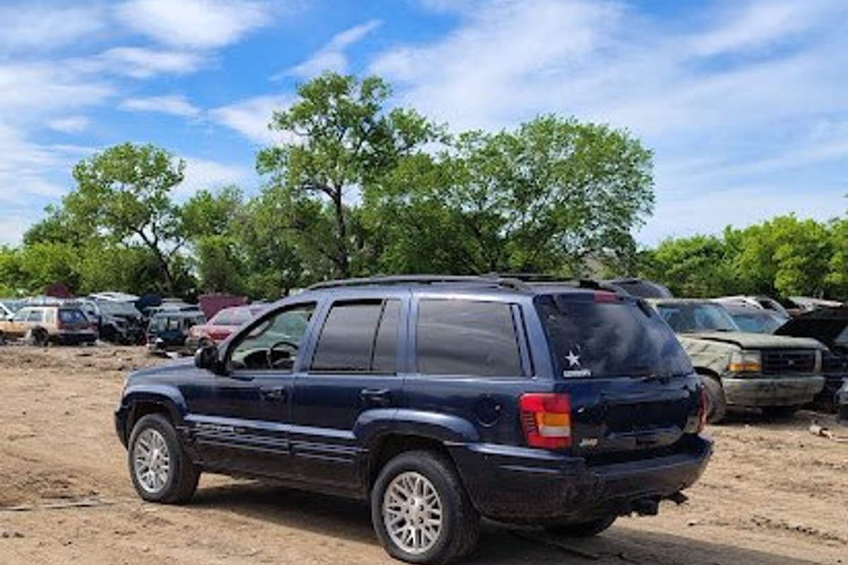 Little John's Wrecking Yard: Dark blue SUV parked on a dirt lot with rows of salvage vehicles and trees in the background under a blue sky