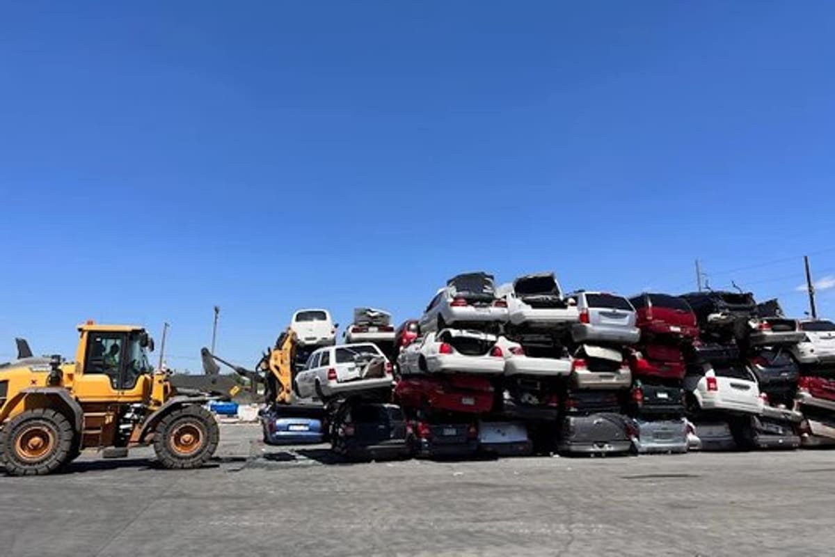 Denver Scrap Metal Recycling: Wide daytime view of stacked crushed cars in a salvage yard with a front loader on paved ground under a clear blue sky