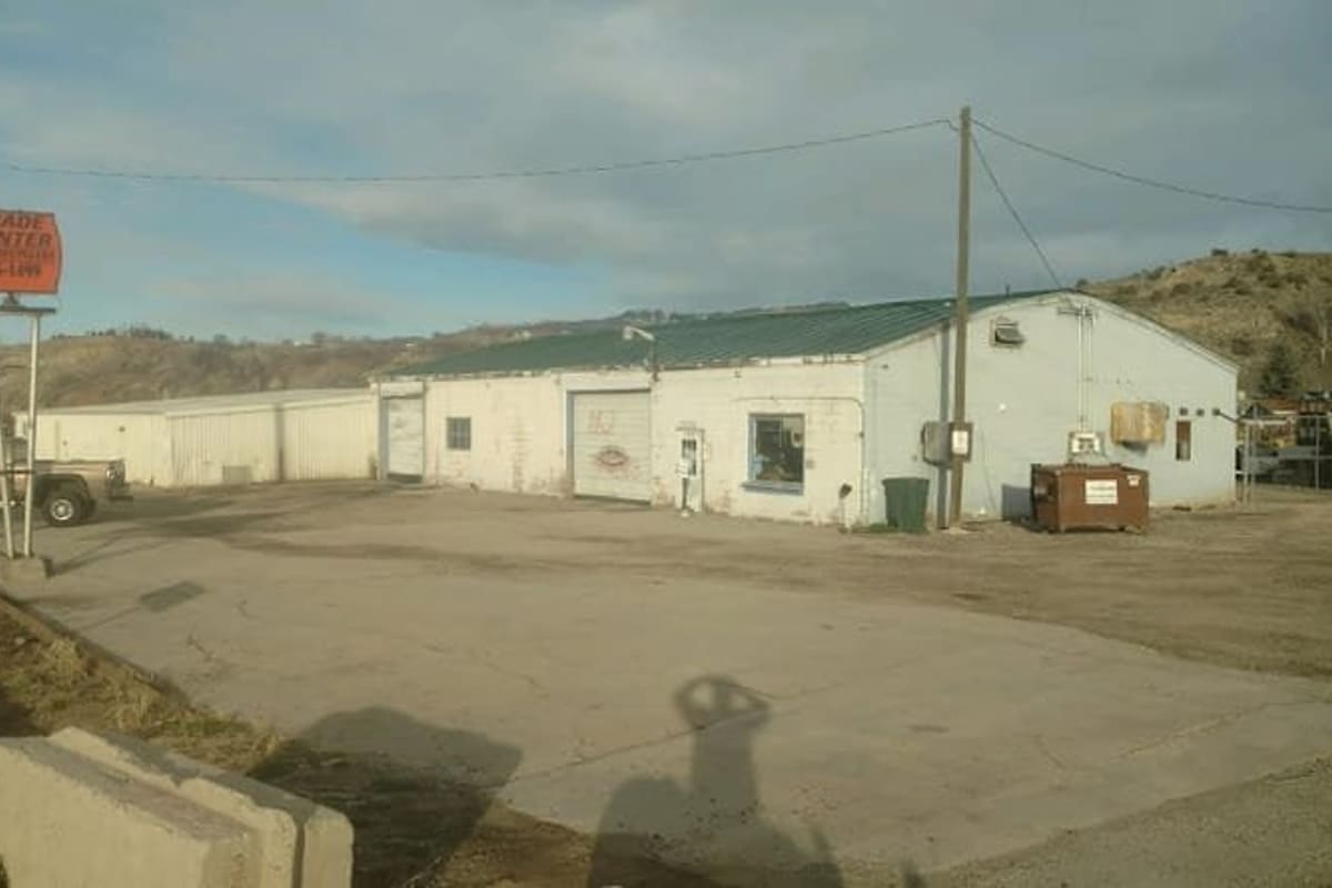 Trade Center Auto Recyclers: Low white metal building with green roof beside a gravel lot and a roadside orange sign on a pole