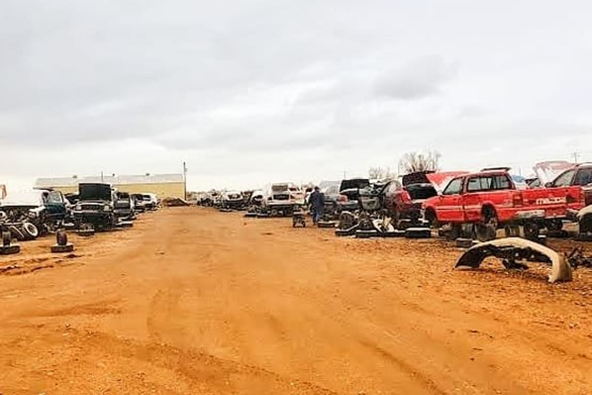Andersens Sales and Salvage 8 St: Dirt lane running between rows of wrecked cars and trucks in a salvage yard under an overcast sky