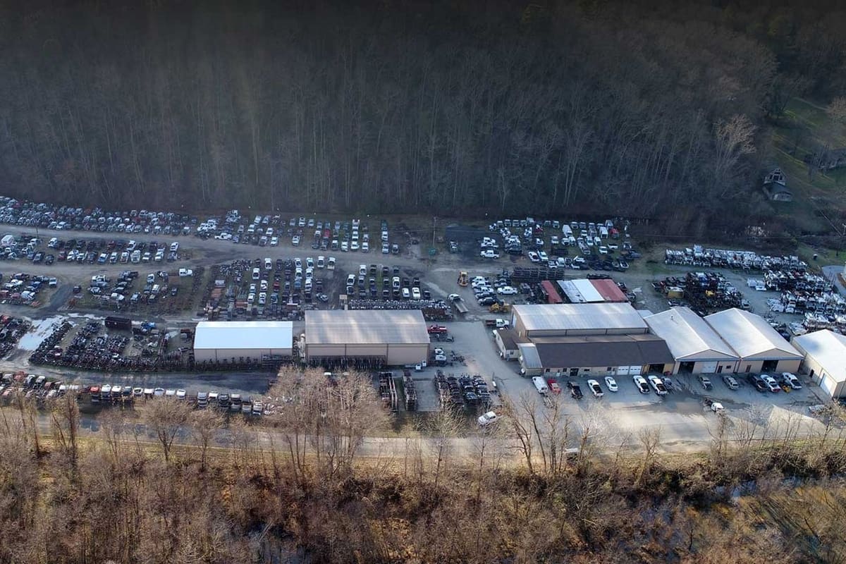 J & T Auto Parts: Aerial view of a large salvage yard with rows of vehicles and several warehouse buildings beside a wooded hillside