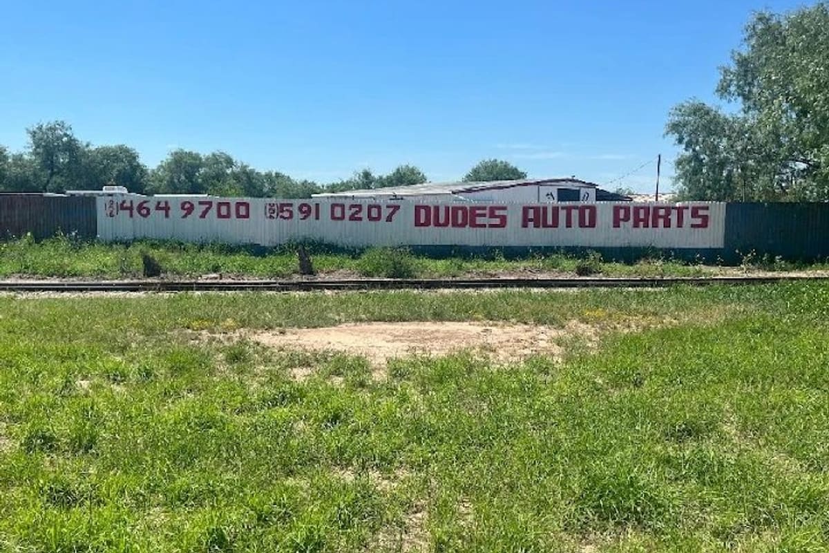 M3 Auto Salvage: Long white fence painted with red lettering and phone numbers beside a grassy lot, with a low building and trees behind