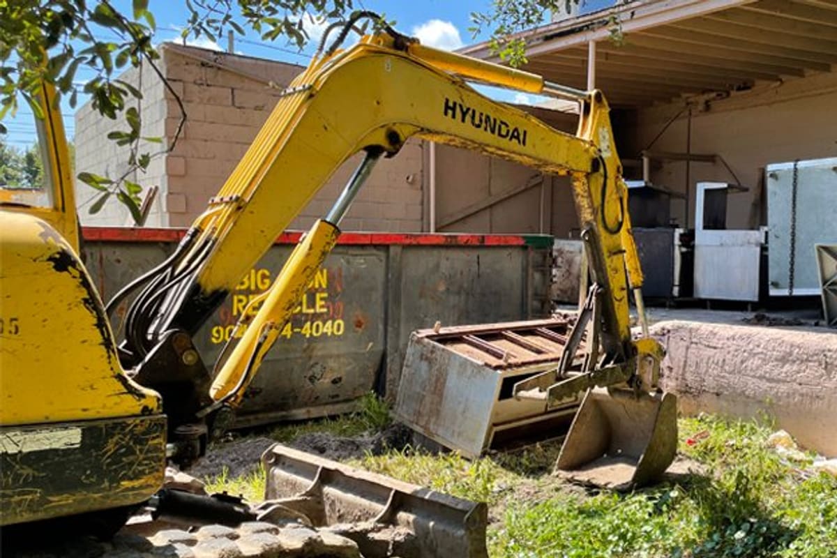 Big Iron Recycling: Yellow excavator working beside a large metal dumpster and a brick building on a recycling lot
