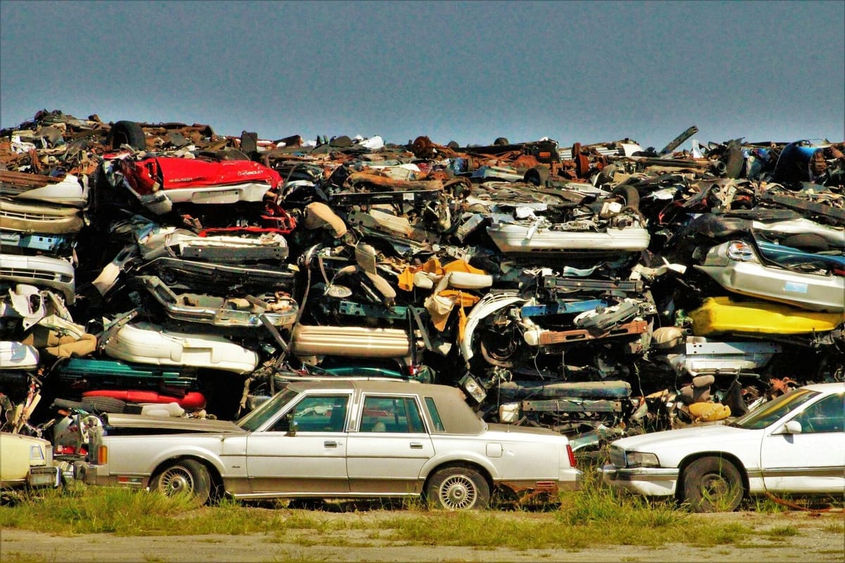 B Auto Recycling: Tall stacks of crushed cars piled behind parked vehicles on a salvage yard lot under a clear sky