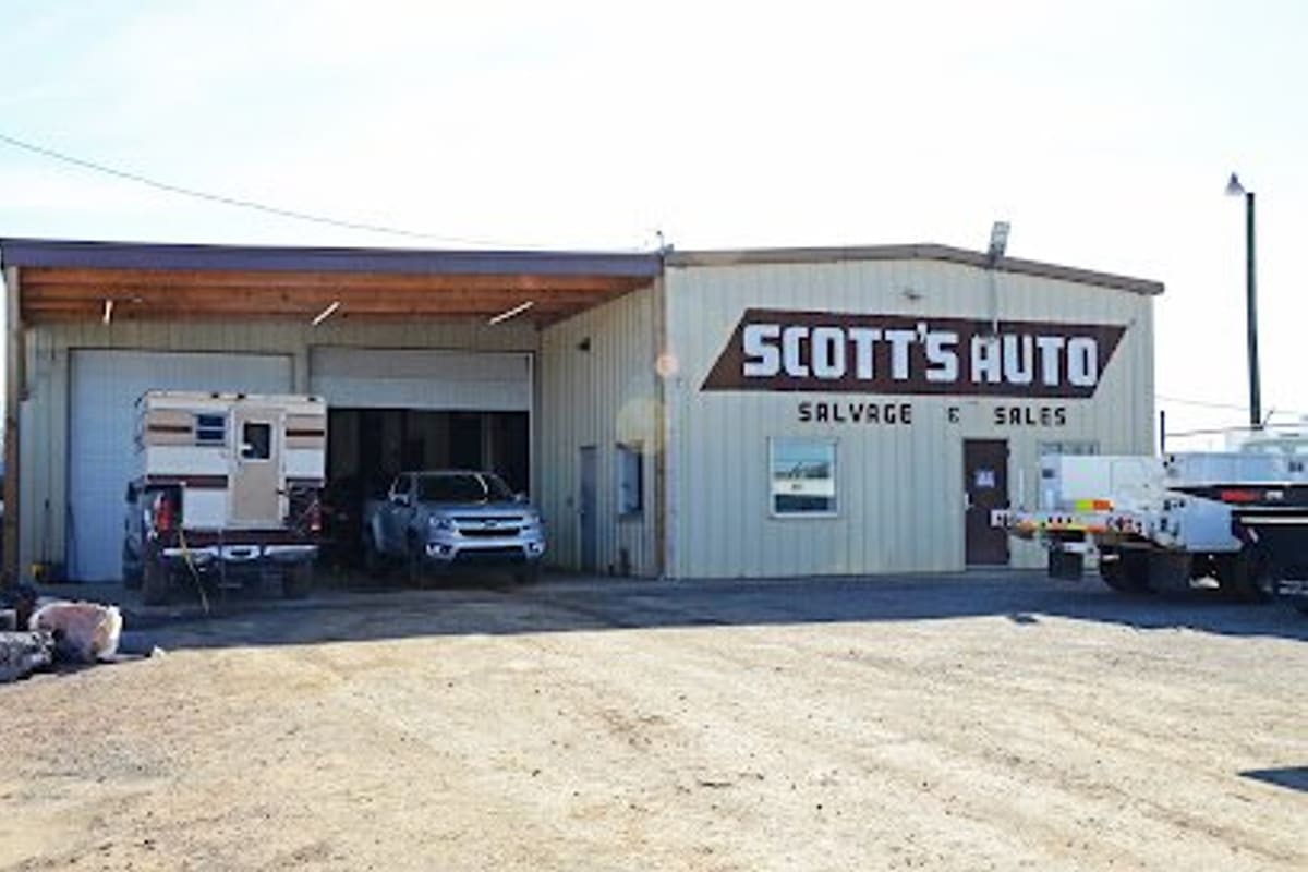 Scott's Auto Salvage and Sales: Tan metal shop building with a large painted sign, open bay with a truck camper and pickup, and a gravel lot in front