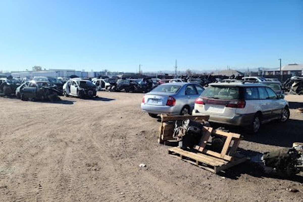 Carbones: Dirt salvage yard with rows of wrecked cars, a silver sedan and wagon in front, and parts on pallets under a clear blue sky