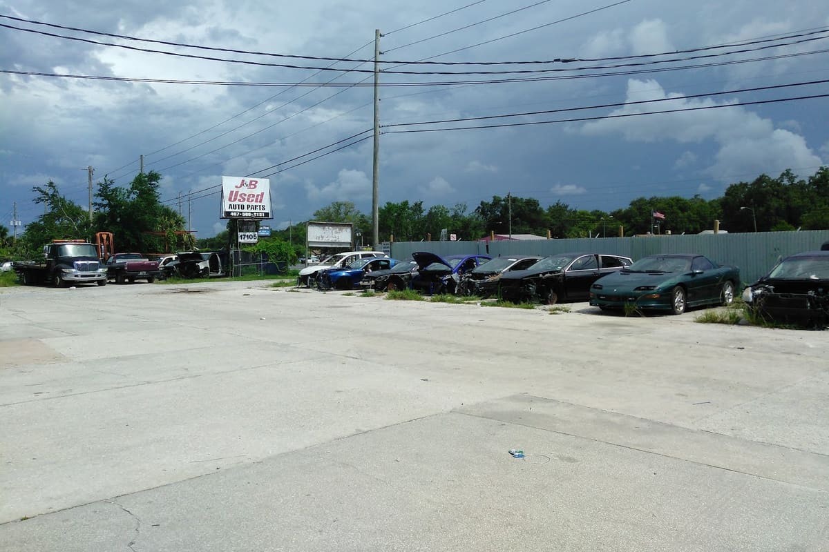 J n B Auto Parts: Wide view of a paved lot with a row of wrecked cars, a tow truck, and a roadside parts sign under a cloudy sky