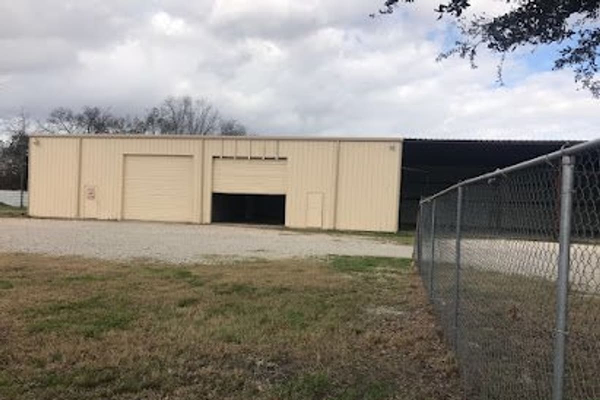 City Auto Recycling: Tan metal warehouse building with two roll-up bay doors beside a gravel lot and a chain-link fence under a cloudy sky