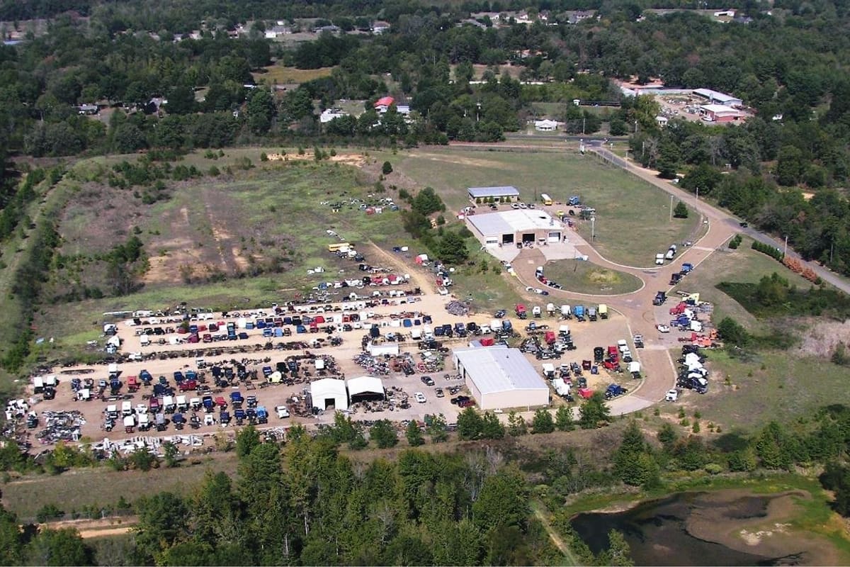 Burtons Heavy Truck Body Shop: Aerial view of a salvage yard with rows of trucks and parts, two metal buildings, and a curved driveway surrounded by trees