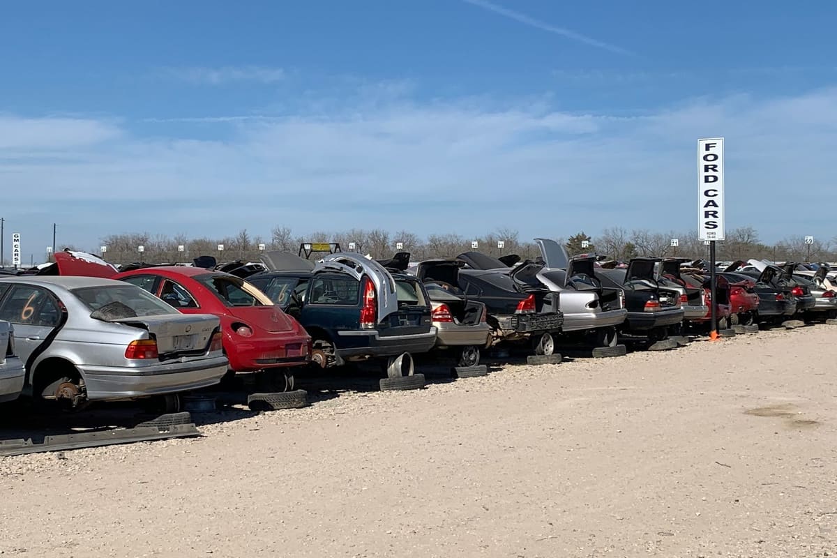 Austin Wrench A Part: Long row of wrecked cars with open hoods and trunks on a gravel lot under a blue sky, with a Ford Cars row sign