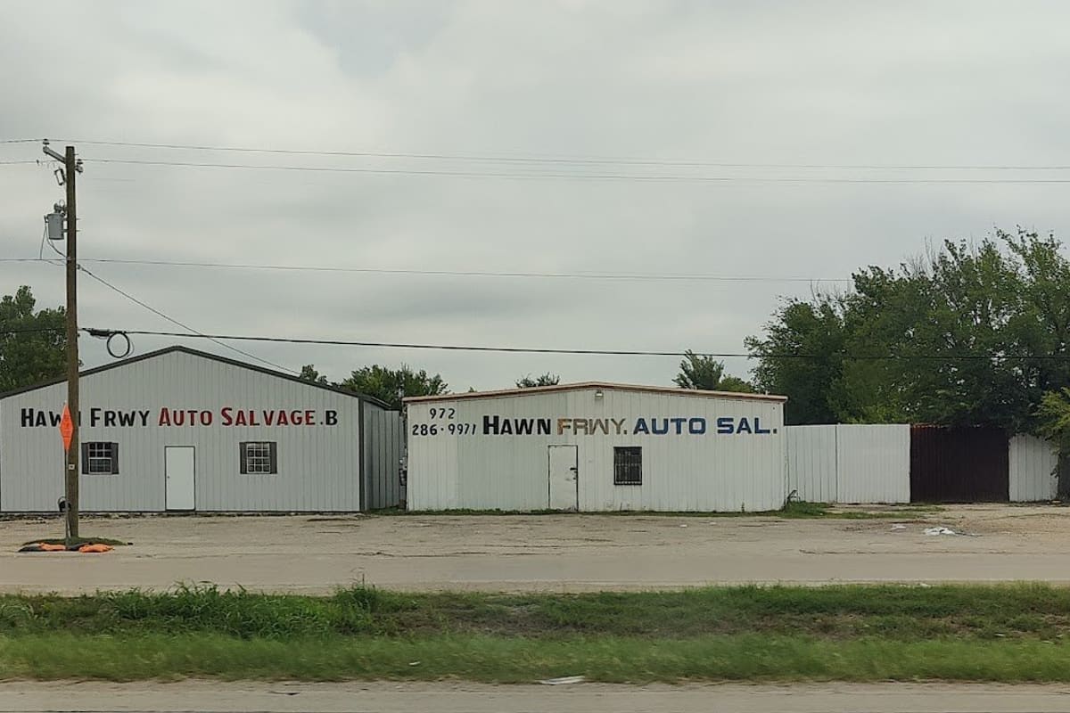 Da Hawn Freeway Auto Salvage: Two white metal buildings with painted signs along a road, with a tall pole sign and trees under an overcast sky