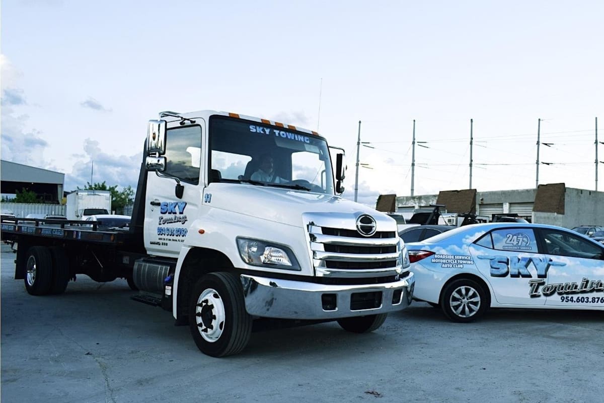 Sky Recycling: Flatbed tow truck and a decal-wrapped car parked on a paved lot with buildings and parked vehicles behind