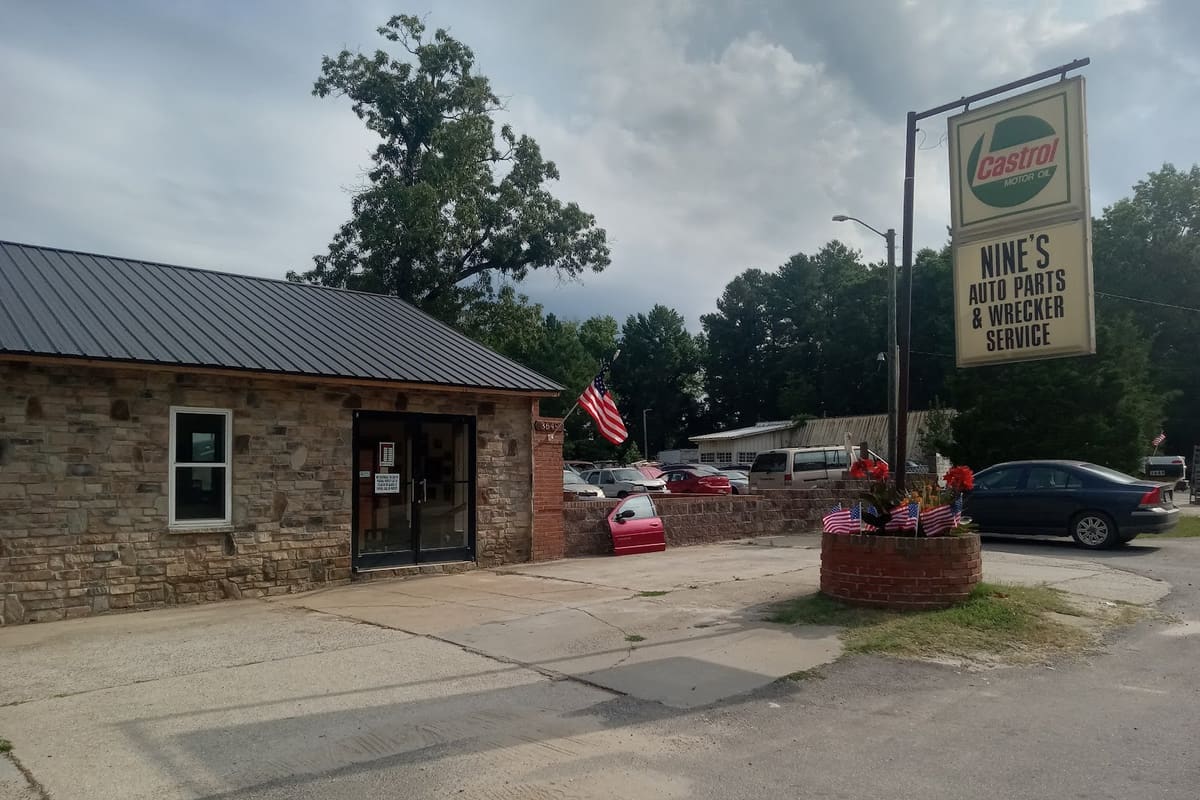 Nine's Auto Parts: Stone office building with metal roof beside a roadside sign for an auto parts and wrecker service, with cars parked behind