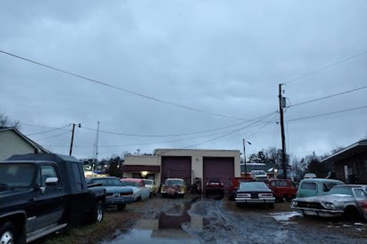 Foothills Salvage: Muddy lot with old cars parked on both sides leading to a two-bay garage building under a cloudy sky