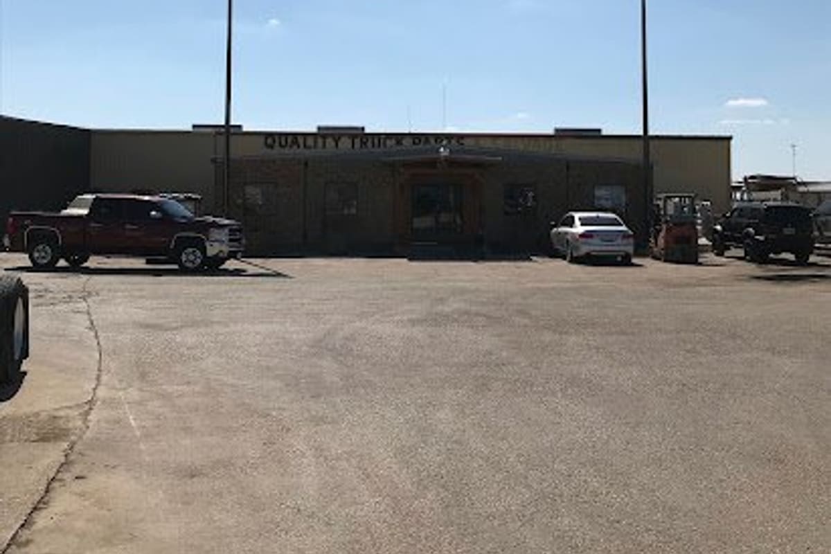 Quality Parts Supply: Single-story parts store with a sign above the entrance, two tall flags, and vehicles parked on a gravel lot under a blue sky