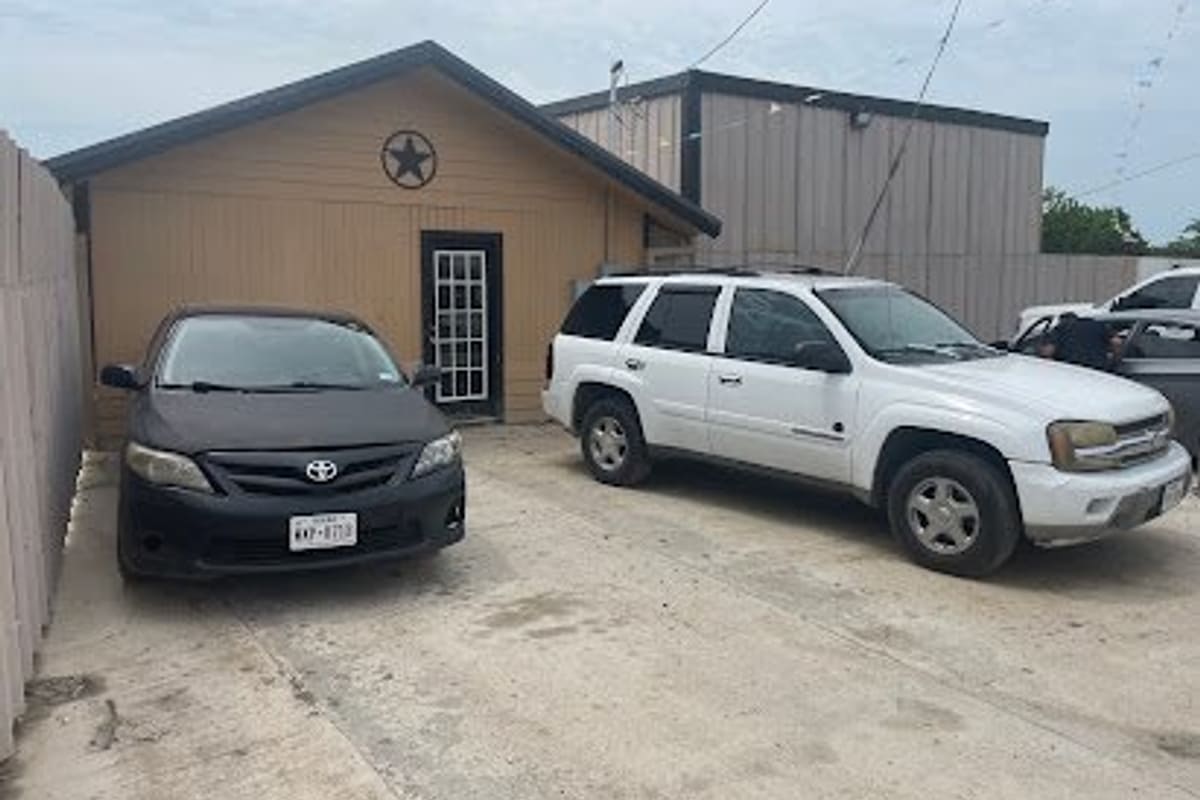 Oz Import & Exotic Auto: Tan shop building with a star on the gable and a glass-paned door, with a black sedan and white SUV parked on a concrete lot
