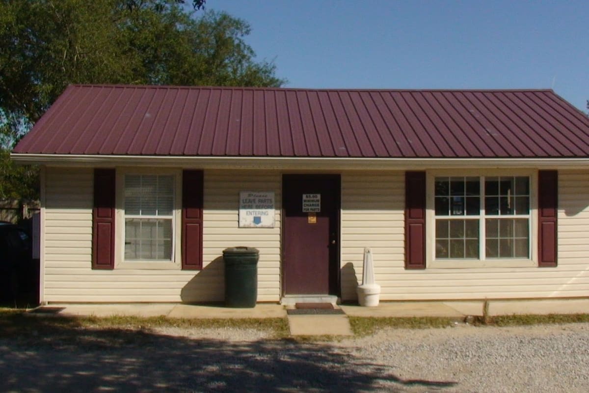 AA Auto Salvage & Recycling: Small tan office building with a maroon metal roof and maroon door, set beside a gravel lot