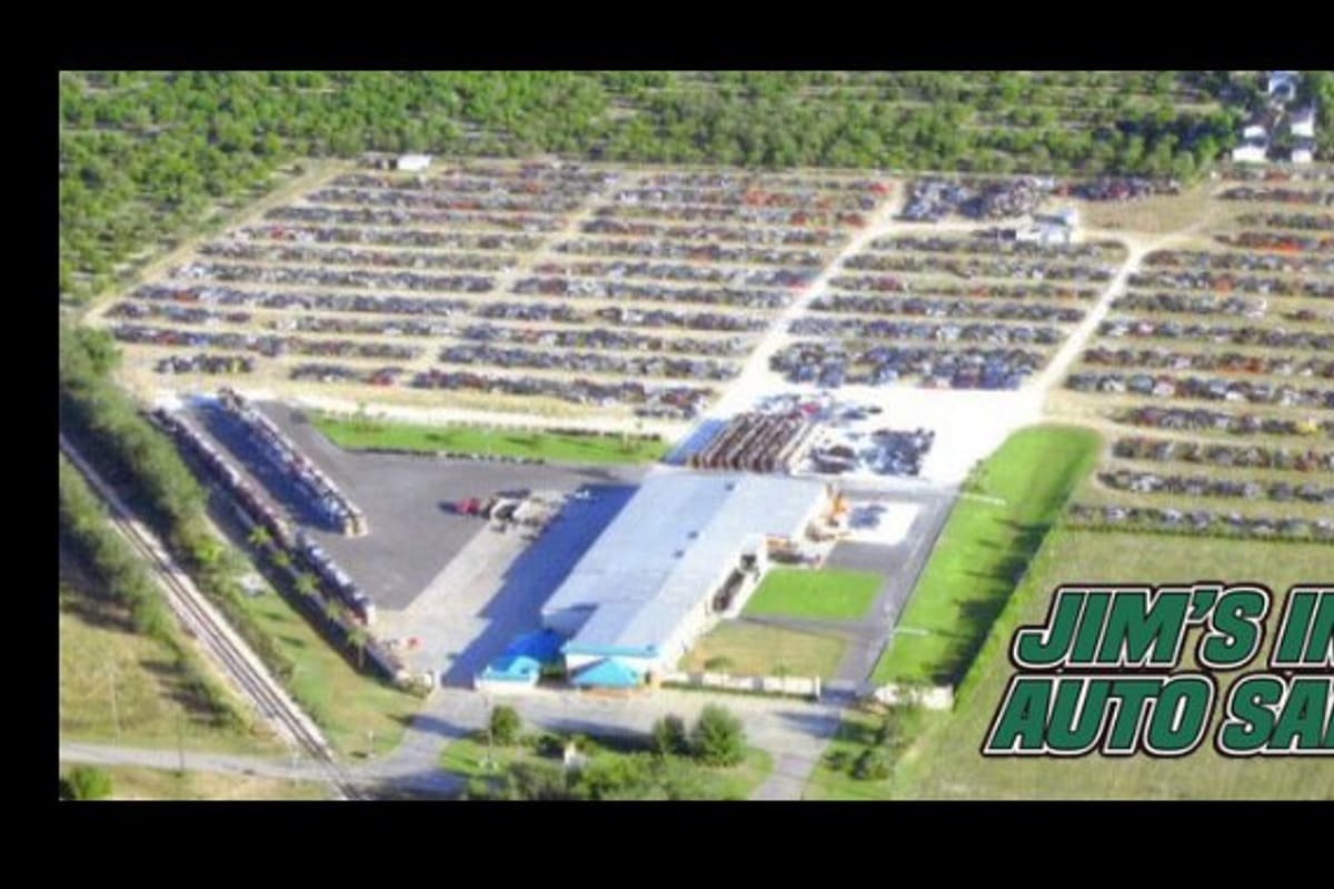 Jim's Import Auto Salvage: Aerial view of a large salvage yard with rows of vehicles, a blue-roofed building and surrounding orchards